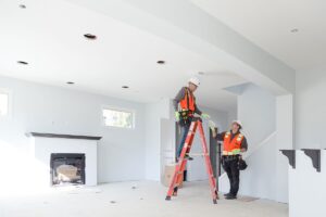 Two electricians in safety gear install ceiling lights in a bright, unfurnished room. One is on a ladder while the other holds tools.