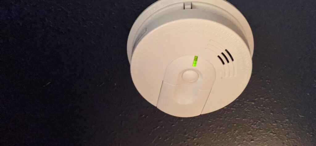 A white smoke detector with a green indicator light, installed by an electrician in Vancouver WA, is mounted on a dark wall.