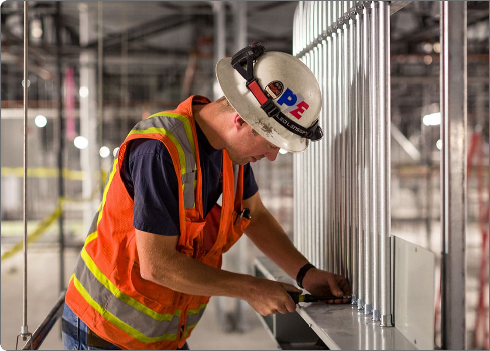 A construction worker wearing a hard hat and safety vest is adjusting metal rods on a structure in an industrial building site, while an electrician inspects the wiring nearby.