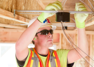 An electrician in a high-visibility vest, hard hat, and safety glasses is installing electrical wiring in a building frame