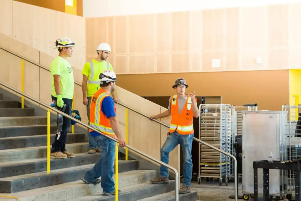 Four construction workers, including an electrician in Vancouver, WA, converse on a stairway at an indoor construction site. They are wearing helmets, safety vests, and work boots.